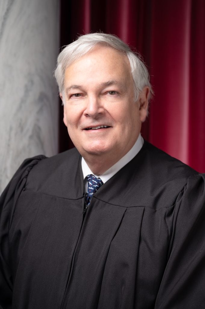 Senior judge in black robe with red curtain background.